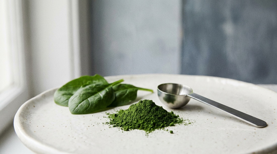 A small pile of deep green powder beside fresh spinach leaves and a steel scoop